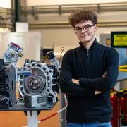 Ein junger Mann mit lockigem Haar und Brille steht neben einem offenen Motor, der auf einem Tisch präsentiert wird. Er trägt einen schwarzen Pullover und hat die Arme verschränkt. Im Hintergrund sind Lehrmaterialien und Stühle sichtbar.