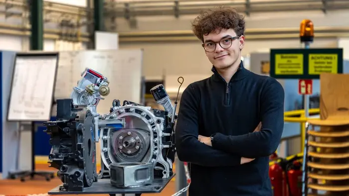 Ein junger Mann mit lockigem Haar und Brille steht neben einem offenen Motor, der auf einem Tisch präsentiert wird. Er trägt einen schwarzen Pullover und hat die Arme verschränkt. Im Hintergrund sind Lehrmaterialien und Stühle sichtbar.