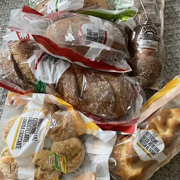 Eine Ansammlung von verschiedenen Brotsorten, verpackt in Plastiktüten. Dazu gehören Brötchen, eine Art Zopf und Kekse. Die Tüten sind teils durchsichtige Folien, die den Inhalt sichtbar machen. Die Brote liegen zufällig auf einem hellen Untergrund.