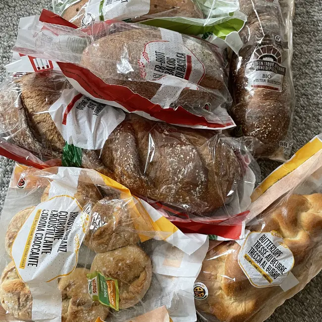 Eine Ansammlung von verschiedenen Brotsorten, verpackt in Plastiktüten. Dazu gehören Brötchen, eine Art Zopf und Kekse. Die Tüten sind teils durchsichtige Folien, die den Inhalt sichtbar machen. Die Brote liegen zufällig auf einem hellen Untergrund.