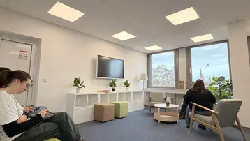 Ein heller Raum mit Teppichboden. Zwei Personen sitzen auf Möbeln: eine auf einer grünen Couch und die andere auf einem Stuhl. An der Wand hängt ein Fernseher. Fenster mit Aussicht auf Pflanzen im Freien. Einige künstliche Pflanzen und kleine Tische sind im Raum verteilt.