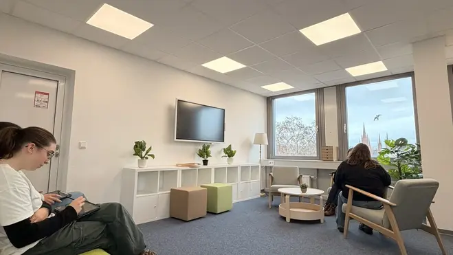 Ein heller Raum mit Teppichboden. Zwei Personen sitzen auf Möbeln: eine auf einer grünen Couch und die andere auf einem Stuhl. An der Wand hängt ein Fernseher. Fenster mit Aussicht auf Pflanzen im Freien. Einige künstliche Pflanzen und kleine Tische sind im Raum verteilt.