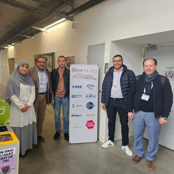 Eine Gruppe von fünf Personen steht in einem Flur. Sie lächeln und posieren für das Foto. Im Hintergrund ist ein Plakat mit verschiedenen Logos und Informationen zum Event „RTPM 2023“ zu sehen.