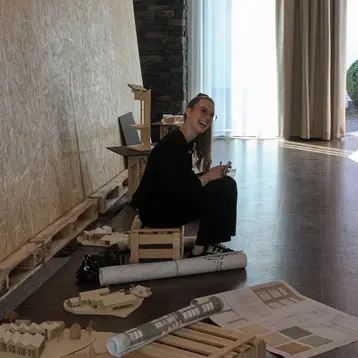 Eine Frau sitzt auf einer kleinen Holzbank in einem modernen Raum. Sie lächelt und hält ein Blatt Papier in der Hand. Im Hintergrund sind große Fenster und eine Wand aus Holzelementen sichtbar. Auf dem Boden liegen Pläne und Zeichnungen.