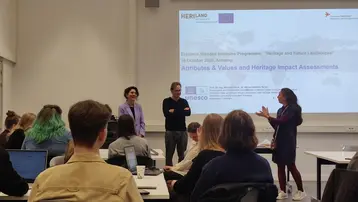 A group of international students are sitting in a seminar room at the University of Antwerp. Two lecturers from HSRM are standing at the front presenting their work on heritage impact assessment, while the participants take notes at their tables. In the background, a projection screen displays the title “Attributes & Values and Heritage Impact Assessments”.