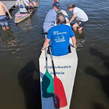 Zwei Männer sitzen auf einem Boot, während eine Frau dabei ist, sich um den Anlegesteg zu kümmern. Im Hintergrund sind weitere Personen, die am Wasser stehen und unterschiedliche Aktivitäten ausführen. Der Mann im Boot trägt ein blaues T-Shirt mit dem Schriftzug „Für große Ideen.“