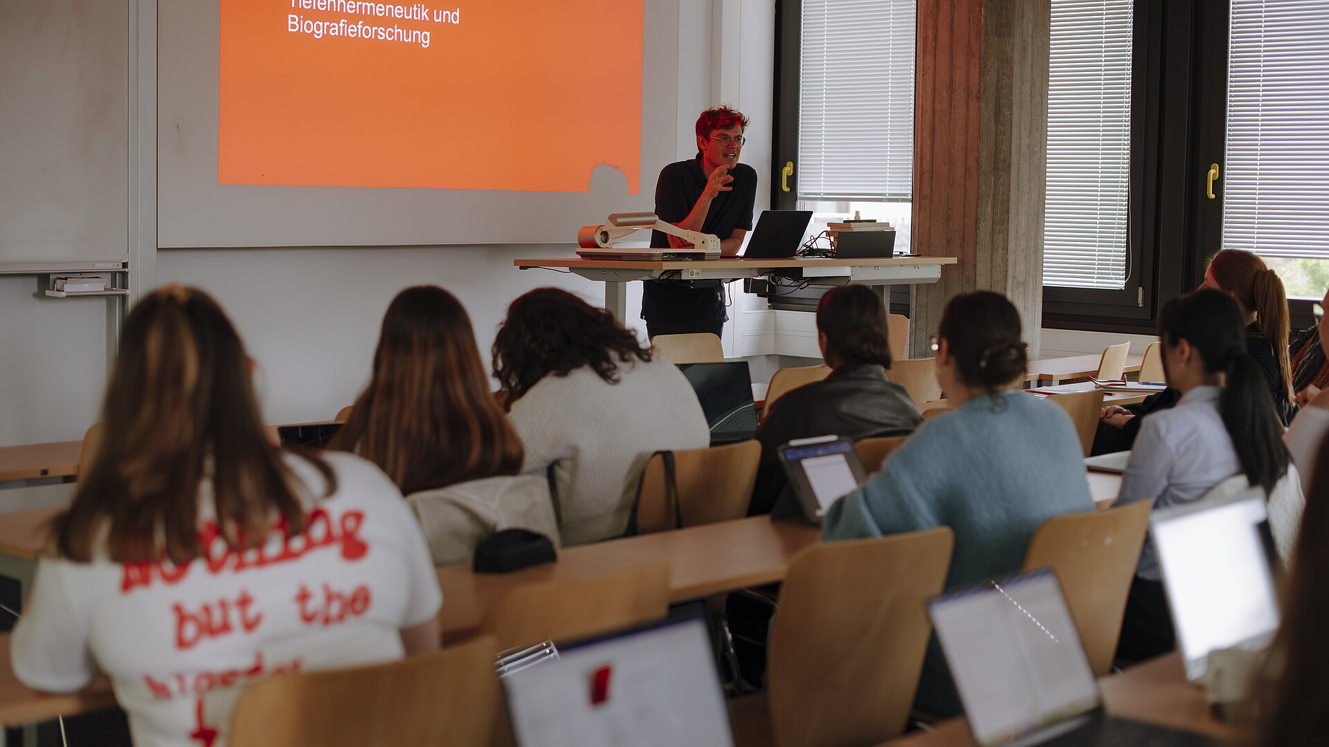 Vortrag KSR Tiefenhermeneutik und Biografieforschung 2 Man sieht einen Vortragenden, der an einem Pult steht und eine Präsentation über "Tiefenhermeneutik und Biografieforschung" hält, so wie das auf seiner Präsentation auf dem großen Bildschirm steht. Studierende sitzen ihm gegenüber an Schreibtischen und hören ihm zu. Sie haben ihre Tablets vor sich.
