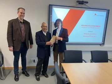 Drei Personen stehen in einem Besprechungsraum. Eine Person überreicht einer anderen ein Dokument oder eine Urkunde. Im Hintergrund ist eine Präsentation auf einem Bildschirm mit dem Titel „Hochschule Riemann“ sichtbar.