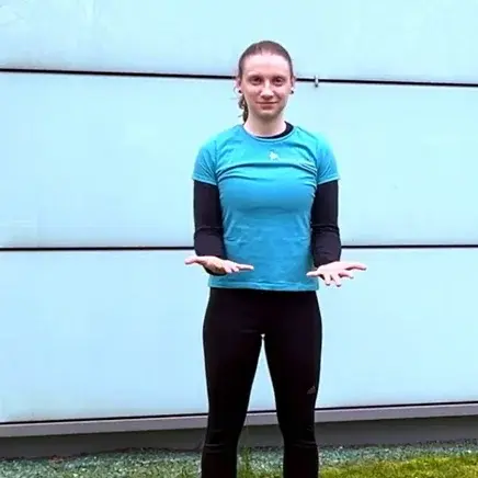 Eine sportliche Person steht in einem blauen Oberteil und schwarzen Leggings vor einer glatten, hellen Wand. Sie hat beide Hände nach vorne ausgestreckt und wirkt freundlich. Die Umgebung ist grün, was eine aktive, gesunde Stimmung vermittelt.