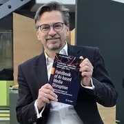 Ein Mann mit Brille hält ein Buch mit dem Titel "Handbook of AI-based Media Disruption" in der Hand. Er sitzt vor einer modernen, minimalistischen Kulisse. Der Fokus liegt auf dem Buch und dem lächelnden Gesicht des Mannes.