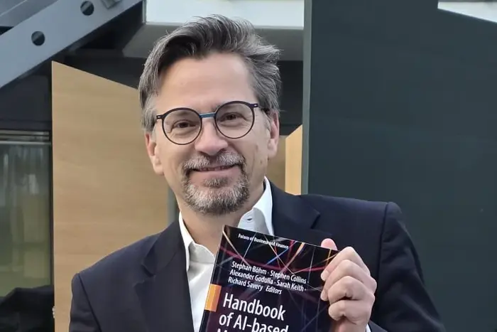 Ein Mann mit Brille hält ein Buch mit dem Titel "Handbook of AI-based Media Disruption" in der Hand. Er sitzt vor einer modernen, minimalistischen Kulisse. Der Fokus liegt auf dem Buch und dem lächelnden Gesicht des Mannes.