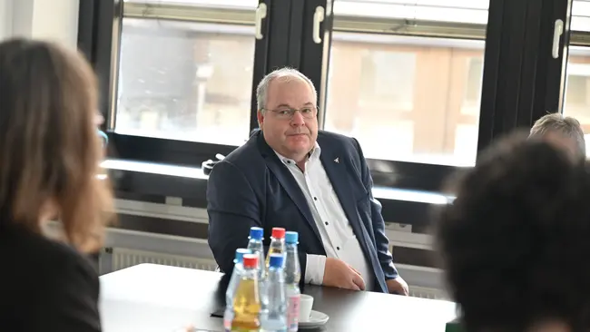 Ein Mann sitzt an einem Tisch in einem Besprechungsraum. Er trägt einen Anzug und schaut in die Runde. Auf dem Tisch stehen mehrere Flaschen mit Getränken. Der Raum hat große Fenster, die Tageslicht hereinlassen.