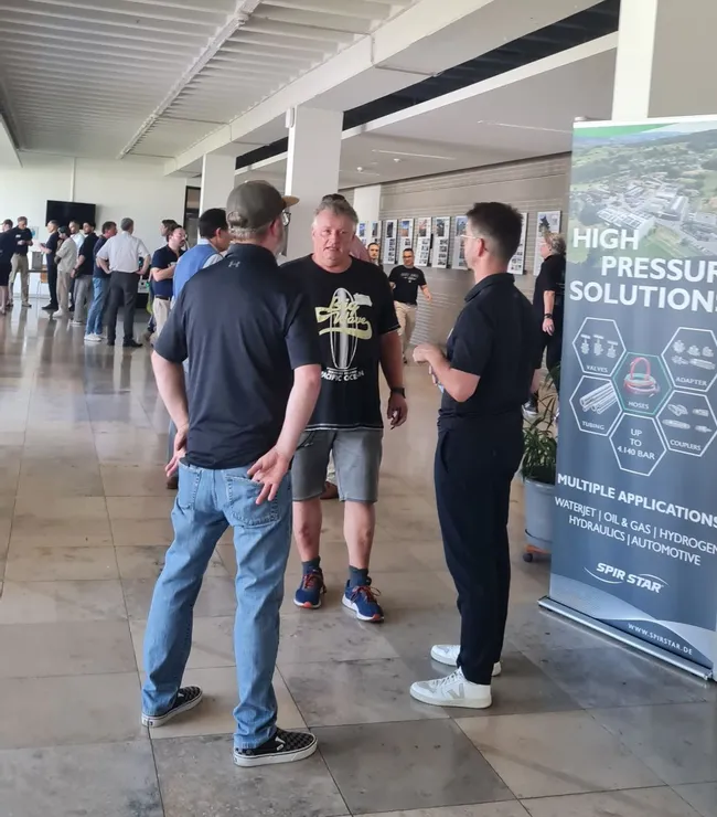 Drei Männer stehen in einem großen, hellen Raum und unterhalten sich. Im Hintergrund sind viele Menschen zu sehen, die ebenfalls miteinander sprechen. Neben den Männern steht ein Stand mit einem Banner, das "High Pressure Solutions" anzeigt.