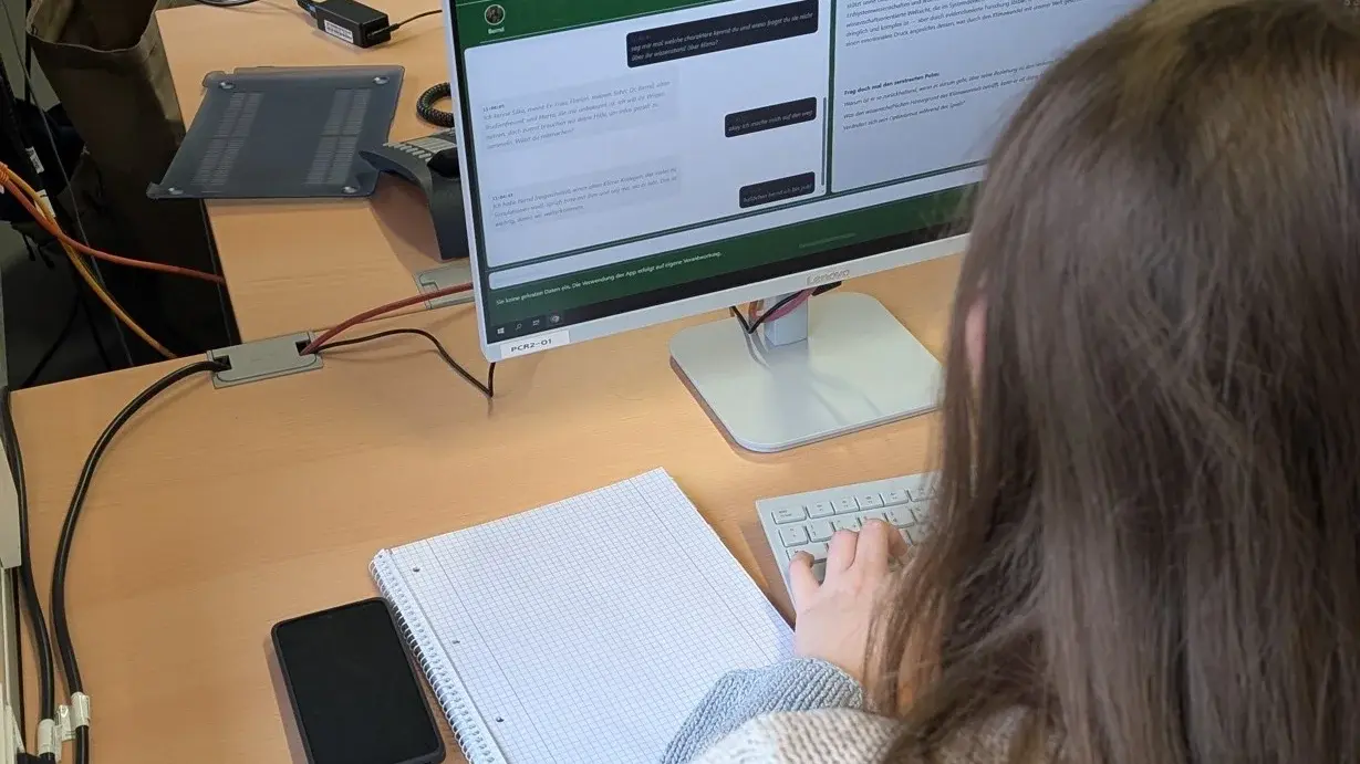Eine Person sitzt an einem Schreibtisch und tippt auf einer Tastatur. Vor ihr steht ein Computerbildschirm, auf dem Text angezeigt wird. Neben dem Computer liegen ein Notizblock und ein Smartphone. Im Hintergrund ist eine grüne Tafel sichtbar.