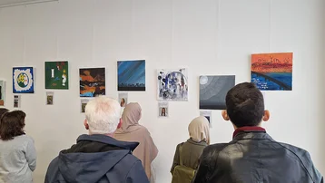 Eine Gruppe von Menschen betrachtet verschiedene Kunstwerke, die an einer weißen Wand hängen. Die Bilder zeigen verschiedene Farben und Themen, während die Betrachter aufmerksam in die Werke schauen.