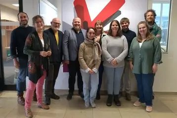 Eine Gruppe von zehn Personen steht in einem Raum vor einem großen Logo mit einem roten V auf weißem Hintergrund. Alle blicken zur Kamera und lächeln. Die Personen sind unterschiedlich gekleidet und scheinen sich in einer professionellen Umgebung zu befinden.