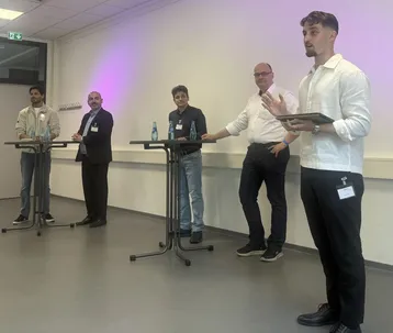 Fünf Männer stehen an Stehtischen in einem Veranstaltungsraum. Alle tragen formelle Kleidung und sind in ein Gespräch vertieft. Einige haben Wasserflaschen auf den Tischen. Die Atmosphäre vermittelt einen professionellen Austausch oder ein Diskussionsforum.