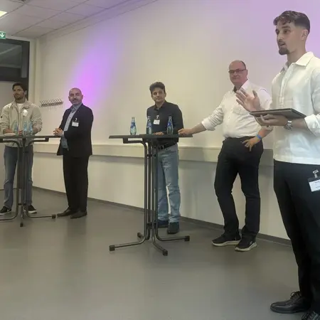Fünf Männer stehen an Stehtischen in einem Veranstaltungsraum. Alle tragen formelle Kleidung und sind in ein Gespräch vertieft. Einige haben Wasserflaschen auf den Tischen. Die Atmosphäre vermittelt einen professionellen Austausch oder ein Diskussionsforum.