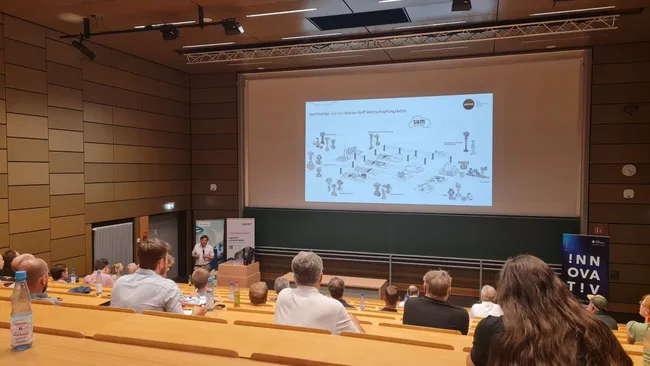 Eine Präsentation in einem großen Vortragssaal. Im Vordergrund steht eine Referentin, die zu einem Diagramm auf einer Leinwand spricht. Im Hintergrund sind zahlreiche Zuhörer auf den Sitzplätzen zu sehen. Die Atmosphäre ist konzentriert und informativ.