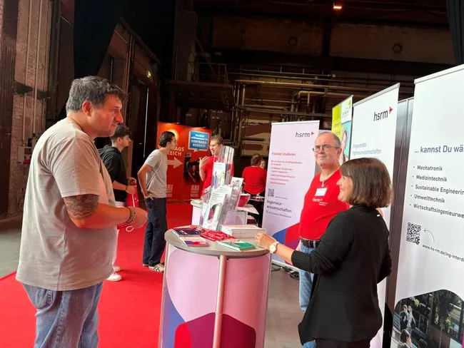 Eine Messe mit mehreren Ständen, an denen Besucher mit Ausstellern sprechen. Im Vordergrund befinden sich eine Frau und ein Mann im Gespräch. Im Hintergrund stehen mehrere andere Personen, die sich ebenfalls austauschen. Die Atmosphäre wirkt informell und einladend.