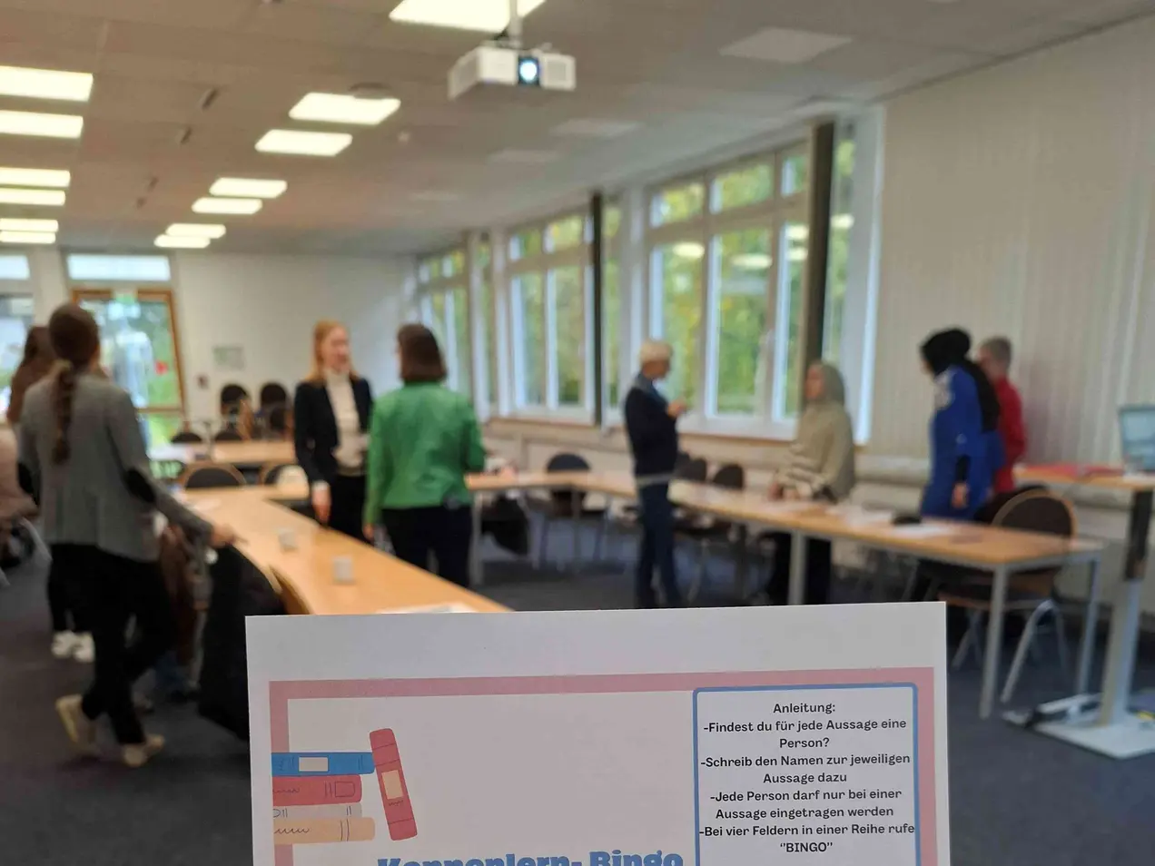 Eine Person hält ein Blatt mit dem Titel "Kennenlern-Bingo" in der Hand. Im Hintergrund sind Menschen in einem hellen Raum, die miteinander interagieren. Das Blatt enthält eine Bingo-Tabelle mit verschiedenen Fragen zum Kennenlernen.
