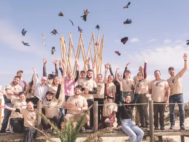 Eine Gruppe von etwa 30 Personen steht auf einer Holzplattform und jubelt. Sie tragen T-Shirts in Beige und lächeln. Einige werfen Handschuhe in die Luft. Im Hintergrund sind Holzstangen zu sehen und der Himmel ist blau mit wenigen Wolken.