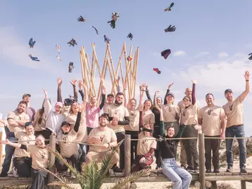 Eine Gruppe von etwa 30 Personen steht auf einer Holzplattform und jubelt. Sie tragen T-Shirts in Beige und lächeln. Einige werfen Handschuhe in die Luft. Im Hintergrund sind Holzstangen zu sehen und der Himmel ist blau mit wenigen Wolken.