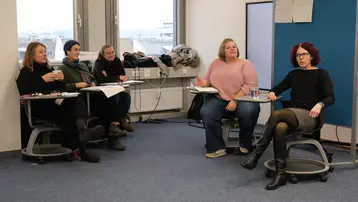 Eine Gruppe von fünf Frauen sitzt in einem Raum, um an einer Diskussion teilzunehmen. Einige halten Notizen oder Getränke in der Hand. Eine Frau spricht, während die anderen aufmerksam zuhören. Der Raum hat große Fenster und einen blauen Akzent in der Dekoration.