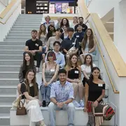 Eine Gruppe von 20 jungen Erwachsenen sitzt und steht auf einer Treppe in einem modernen Gebäude. Alle tragen Namensschilder. Die Atmosphäre ist freundlich und entspannt, und die Personen lächeln oder schauen in die Kamera.