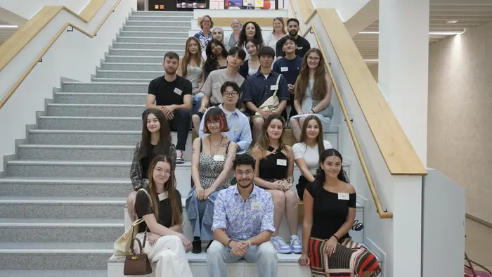 Eine Gruppe von 20 jungen Erwachsenen sitzt und steht auf einer Treppe in einem modernen Gebäude. Alle tragen Namensschilder. Die Atmosphäre ist freundlich und entspannt, und die Personen lächeln oder schauen in die Kamera.