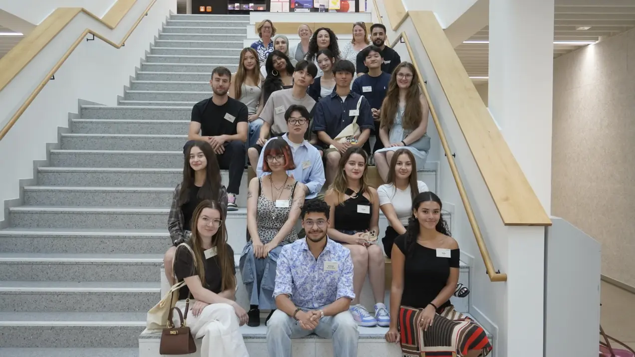 Eine Gruppe von 20 jungen Erwachsenen sitzt und steht auf einer Treppe in einem modernen Gebäude. Alle tragen Namensschilder. Die Atmosphäre ist freundlich und entspannt, und die Personen lächeln oder schauen in die Kamera.