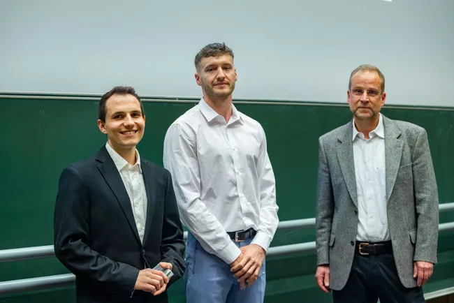 Drei Personen stehen in einem Vortragssaal vor einer grünen Wand. Zwei Männer tragen formelle Kleidung, während der dritte in einem hellen Hemd und hellen Hosen gekleidet ist. Alle drei lächeln und scheinen an einem Event oder einer Präsentation teilzunehmen.