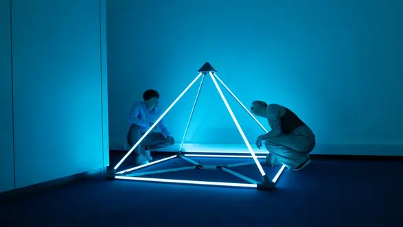 Zwei Personen untersuchen eine leuchtende, pyramidenförmige Skulptur aus Neonrohren in einem blauen Raum. Die Skulptur strahlt helles Blau aus und steht auf dem Boden, während die Personen in unterschiedlichen Positionen darauf fokussiert sind.