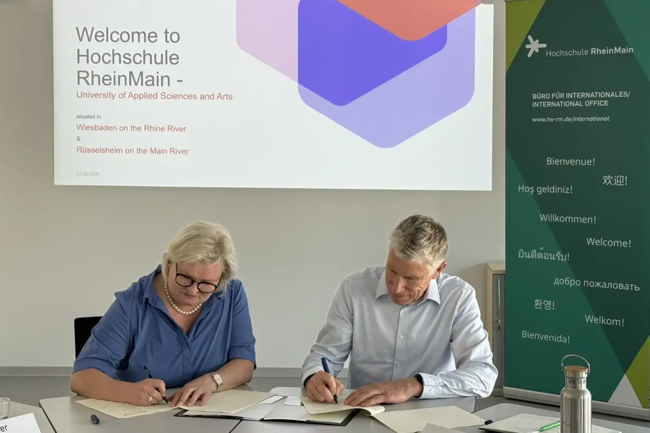 Eine Frau und ein Mann sitzen an einem Tisch und schreiben. Im Hintergrund befindet sich ein Bildschirm mit der Aufschrift "Welcome to Hochschule RheinMain" sowie Informationen zur Universität und zur Website. Rechts steht ein Banner mit mehrsprachigen Grußformeln.