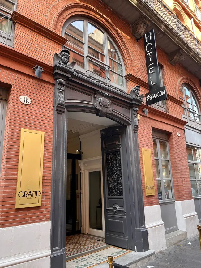Der Eingang zum Hotel Le Grand Balcon zeigt eine elegante, verzierten Tür mit einem stilvollen schwarzen Rahmen. Über der Tür hängt ein Schild mit der Aufschrift "HOTEL". Die Fassade ist aus roten Ziegeln und vermittelt einen historischen Charme.