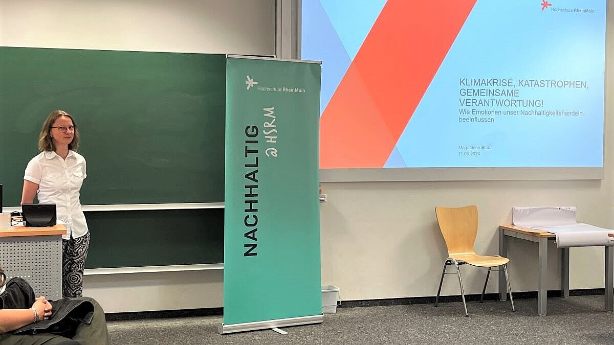 Magdalene Biada steht vor einer Tafel, neben der ein Banner und an der Wand eine Präsentation zu sehen sind (Text: "Nachhaltig @ HSRM", "Klimakrise, Katastrophen, gemeinsame Verantwortung"