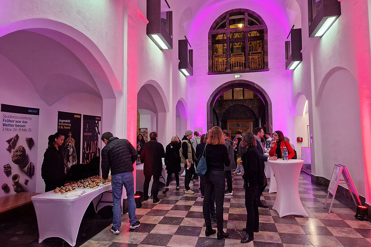 Das Foyer des Museums Wiesbaden erstrahlt in rotem und pinken Licht. An Stehtischen stehen die Besucher:innen mit Getränken und Snacks. 