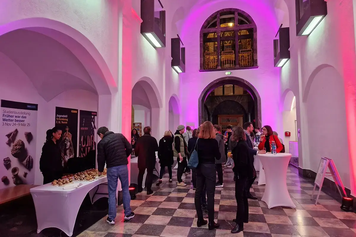 Das Foyer des Museums Wiesbaden erstrahlt in rotem und pinken Licht. An Stehtischen stehen die Besucher:innen mit Getränken und Snacks. 