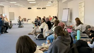 Eine Gruppe von etwa 20 Personen sitzt in einem Seminarraum. Sie hören einer Präsentation zu, während einige Notizen machen. Der Raum ist hell, mit großen Fenstern und einem Flipchart. Die Teilnehmenden sind unterschiedlich gekleidet und scheinen aufmerksam zu sein.