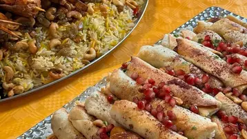 Auf dem Bild sind zwei Gerichte zu sehen: Auf der linken Seite ein großes Blech mit gelbem Reis und Meeresfrüchten, garniert mit Cashewkernen. Rechts daneben ist ein flaches Blech mit gefüllten Teigrollen, dekoriert mit Granatapfelkernen und Gewürzen. Der Hintergrund ist orangefarben.