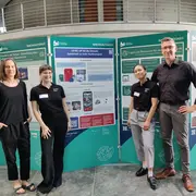 Vier Personen stehen vor mehreren Informationspostern zum Thema Nachhaltigkeit. Sie tragen schwarze T-Shirts mit Namensschildern und stehen in einer modernen Umgebung. Die Poster präsentieren verschiedene Aspekte von nachhaltiger Entwicklung und Technologien.