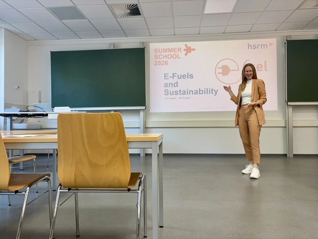 Eine Frau steht in einem Klassenraum und präsentiert ein Thema über E-Fuels und Nachhaltigkeit. Hinter ihr ist ein großes Plakat mit dem Titel „E-Fuels and Sustainability“ und der Jahreszahl 2026 zu sehen. Der Raum ist modern und schlicht eingerichtet.