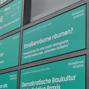 Der Bildinhalt zeigt mehrere grüne Plakatwände mit Texten. Themen umfassen einen Workshop zum #DtD26, die Frage nach der Nutzung von Straßenräumen, und Diskussionen zur Baukultur und sozialen Transformationen. Das Datum und der Titel einer Veranstaltung sind ebenfalls erwähnt.