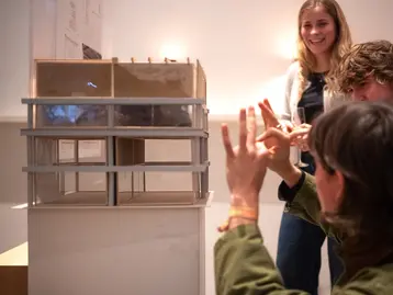 Three people are standing around an architectural model. One person is pointing at the model with their hands, while the others are watching with interest and smiling.