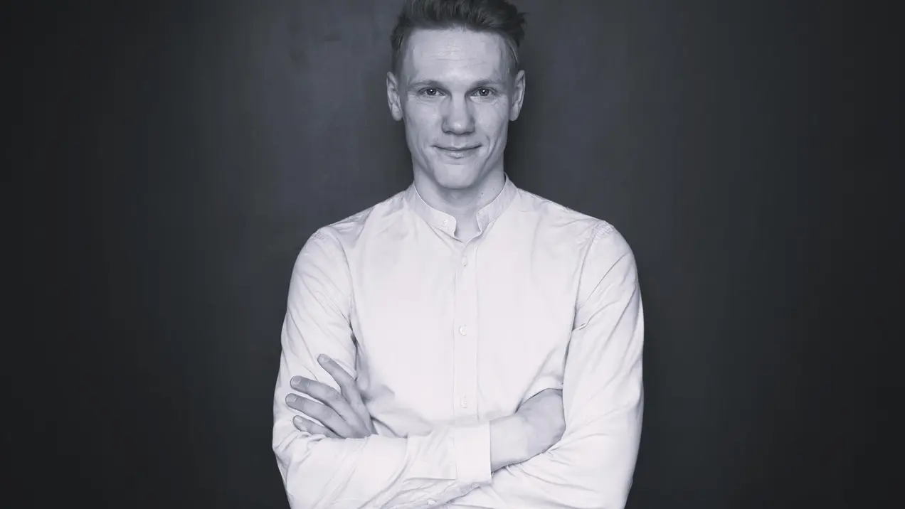 Ein Mann mit kurzem Haar steht vor einem dunklen Hintergrund. Er trägt ein weißes Hemd und hat die Arme vor der Brust verschränkt. Der Gesichtsausdruck ist freundlich und selbstbewusst.
