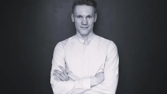 Ein Mann mit kurzem Haar steht vor einem dunklen Hintergrund. Er trägt ein weißes Hemd und hat die Arme vor der Brust verschränkt. Der Gesichtsausdruck ist freundlich und selbstbewusst.