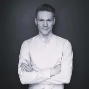 Ein Mann mit kurzem Haar steht vor einem dunklen Hintergrund. Er trägt ein weißes Hemd und hat die Arme vor der Brust verschränkt. Der Gesichtsausdruck ist freundlich und selbstbewusst.