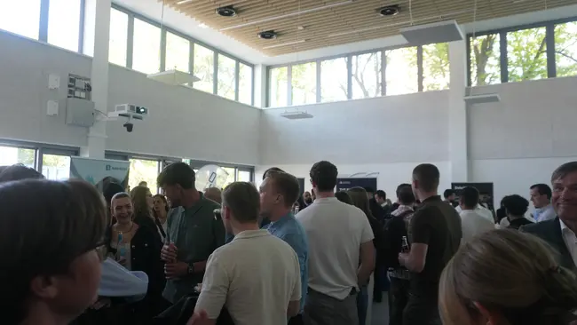 Eine Gruppe von Menschen steht in einem hellen Raum mit großen Fenstern. Die Anwesenden unterhalten sich, halten Getränke in der Hand und wirken in geselliger Stimmung. Der Raum hat holzverkleidete Decken und eine moderne Ausstattung.