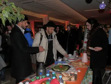 Eine Gruppe von Menschen steht an einem Tisch, der mit Getränken und Snacks gedeckt ist. Auf dem Tisch sind verschiedene Speisen, darunter Salat und Gebäck. Die Atmosphäre wirkt gesellig, und einige Personen unterhalten sich, während sie die bereitgestellten Speisen betrachten.