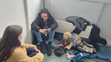 Eine Frau spricht mit einem Mann, der auf dem Boden sitzt und scheinbar in einer schwierigen Lebenssituation ist. Neben ihm liegt ein Teddybär und zerknüllte Kleidung. Die Atmosphäre wirkt ernst und bedürftig.